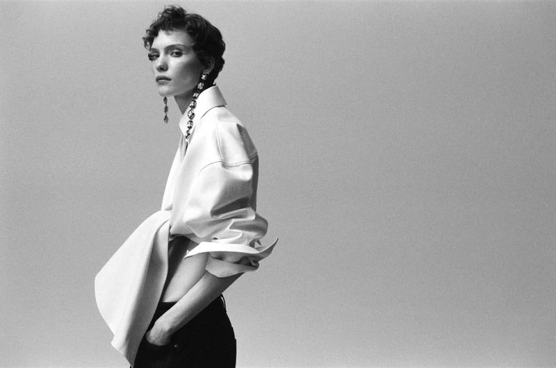 A woman poses indoors wearing a white blouse with large sleeves and dark pants against a light grey background in a studio shot.