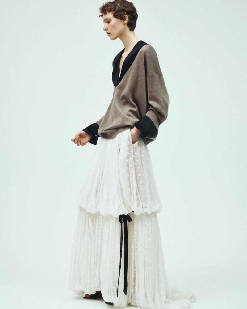 A young woman stands in a studio wearing a grey sweater with a black collar and a white tiered skirt with a black ribbon.