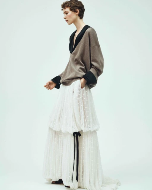 A young woman stands in a studio wearing a grey sweater with a black collar and a white tiered skirt with a black ribbon.