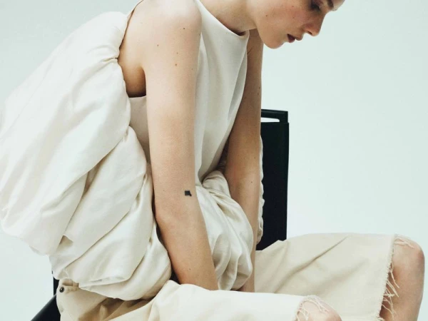 Fashion photography of a seated, fair-skinned woman in an oversized cream dress against a clean white backdrop, with a black chair.