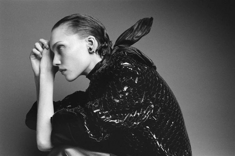 Black and white fashion portrait of a seated model in a high-necked, sequined top, with her hands resting on her face in profile.