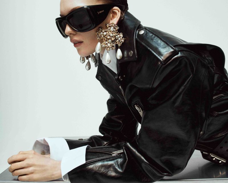 Woman wearing sunglasses, black leather jacket, white collared shirt, and oversized floral earrings, posing in a studio environment.