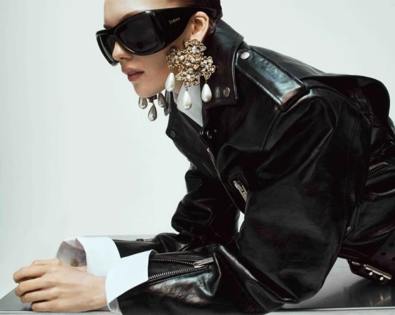Woman wearing sunglasses, black leather jacket, white collared shirt, and oversized floral earrings, posing in a studio environment. 