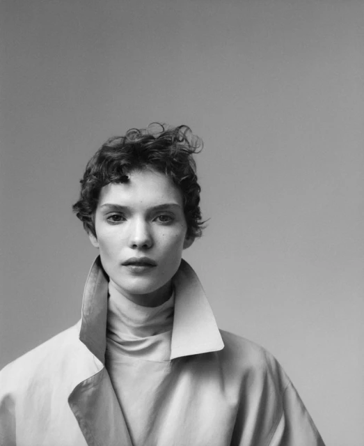 Black and white portrait of an adult woman with short curly hair wearing a trench coat and turtleneck sweater in a photo studio.