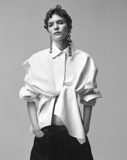 Monochrome studio portrait of a woman in a white button-down shirt and dark pants, accessorized with long, geometric earrings.