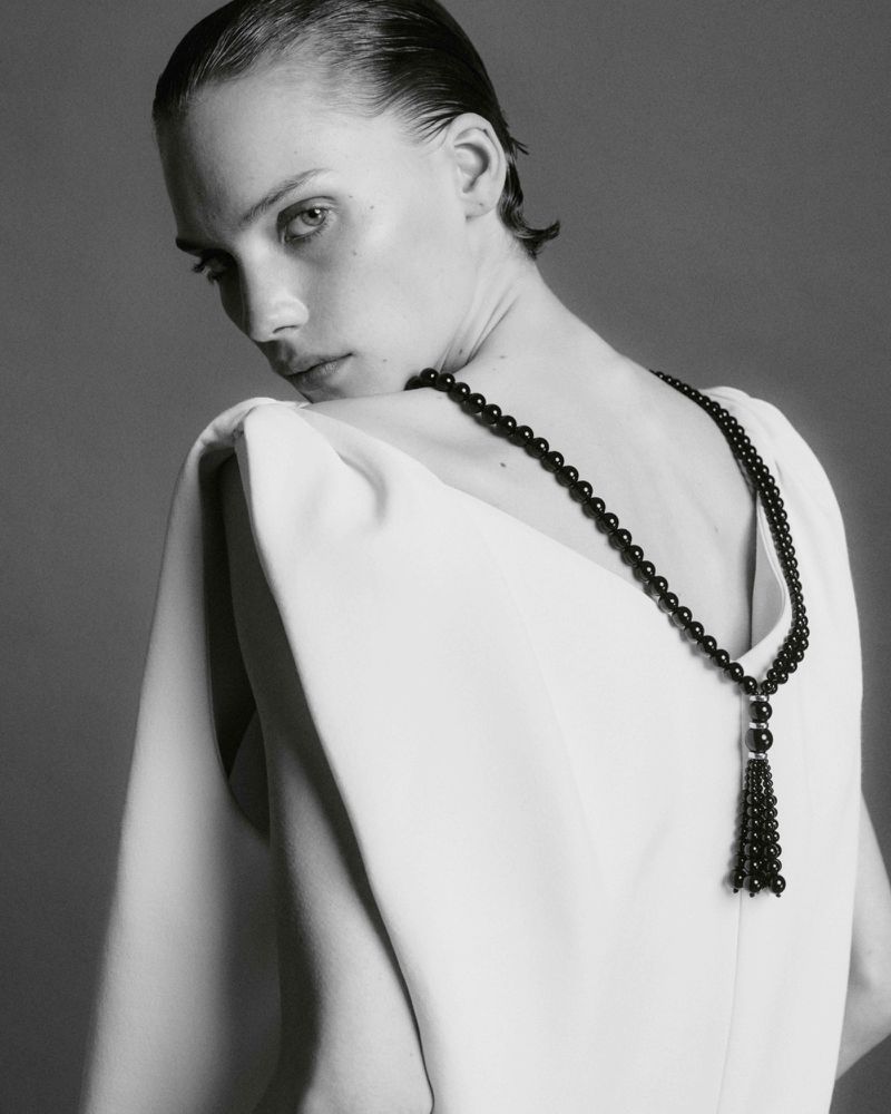Monochrome portrait of a woman in an elegant white dress and black beaded tassel necklace against a solid grey backdrop, fashion photography.