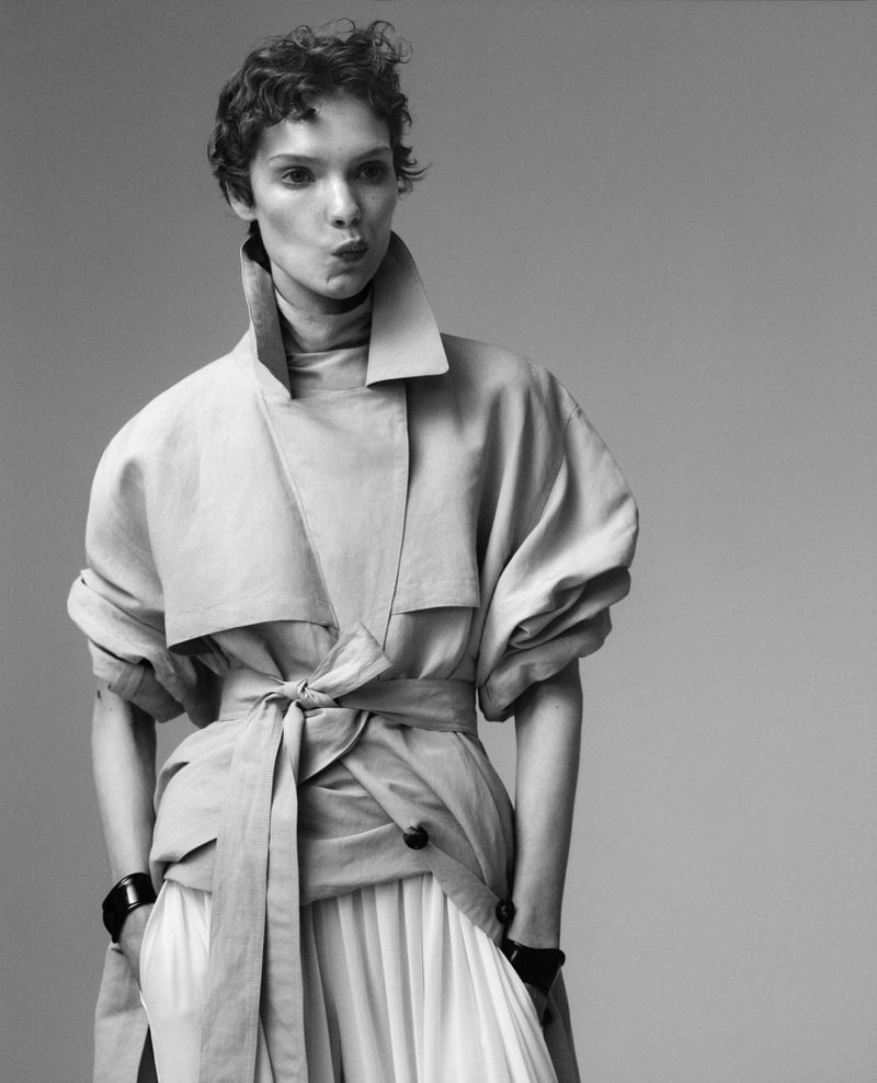 Black and white fashion shot of a female model with short curly hair wearing a light-colored trench coat and skirt in a studio setting.