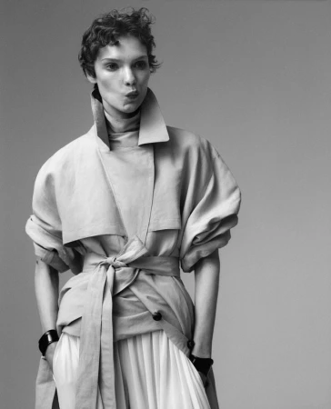 Black and white fashion shot of a female model with short curly hair wearing a light-colored trench coat and skirt in a studio setting.