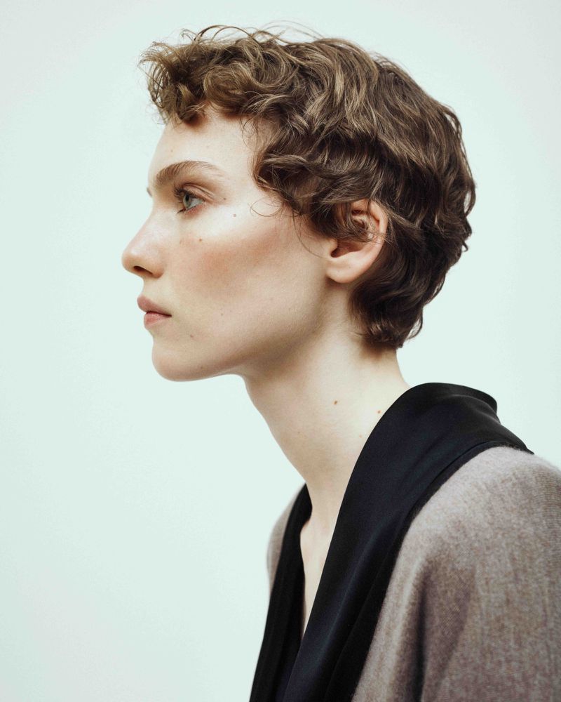 Portrait of a woman with curly brown hair in profile against a neutral background, wearing a black top with a brown cardigan.