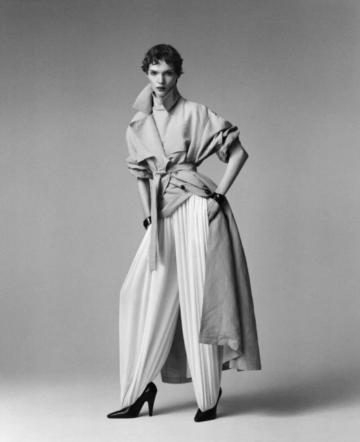 Fashion shot of a woman in a belted trench coat and wide-legged pleated pants, striking a pose against a light grey backdrop.