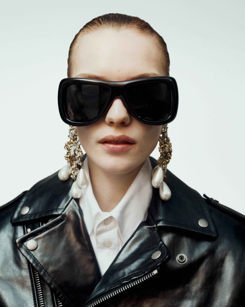 Fashionable woman in a black leather jacket and oversized sunglasses, accessorized with statement pearl earrings, against a white background.