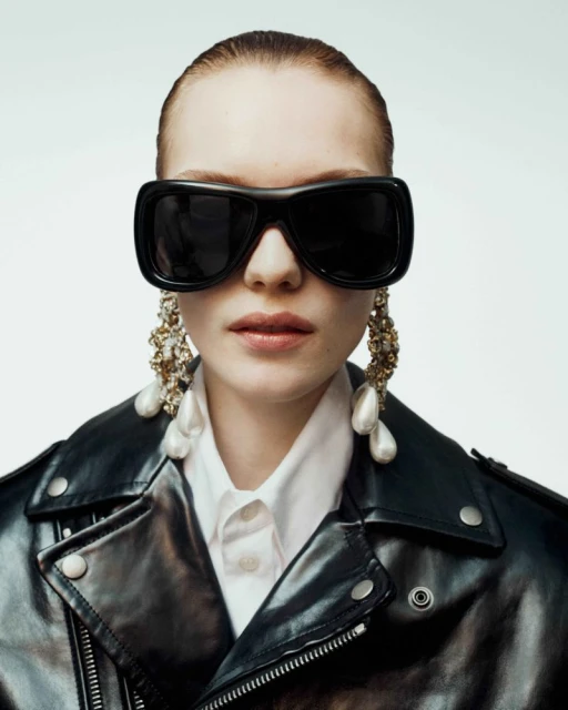 Fashionable woman in a black leather jacket and oversized sunglasses, accessorized with statement pearl earrings, against a white background.