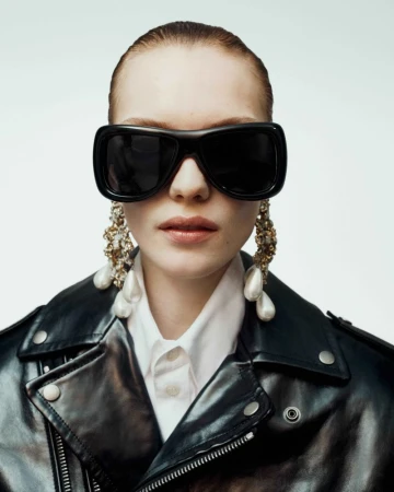 Fashionable woman in a black leather jacket and oversized sunglasses, accessorized with statement pearl earrings, against a white background.