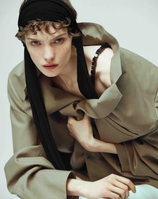 Portrait of a young adult woman with a black headscarf and an oversized tan jacket posing in a studio with a neutral background.