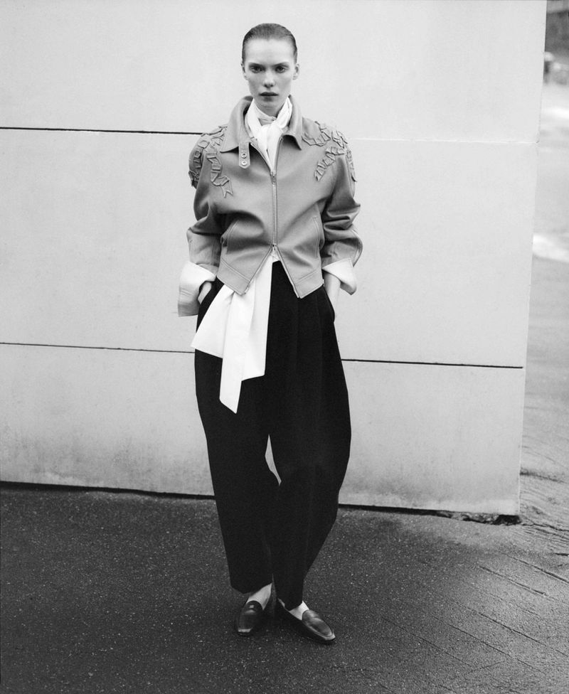 Full shot of a slender adult woman standing outdoors, wearing a leather jacket with shoulder ornamentation, black pants and flats.