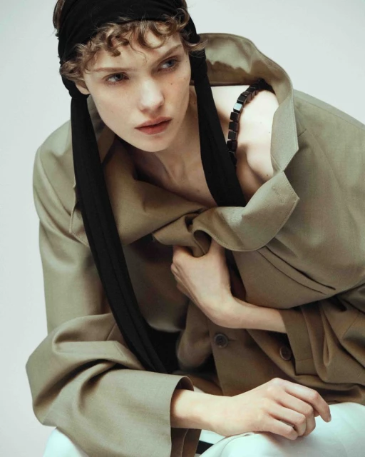 Portrait of a young woman with curly hair wearing a black headscarf and an oversized beige jacket against a white background.