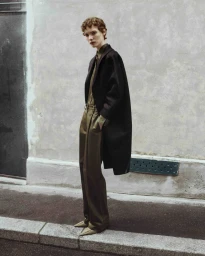Model Penelope Ternes posing in an oversized black coat and olive trousers on a street curb against a pale textured wall.