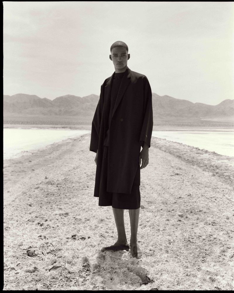 Model Samuel Elie stands in an oversized black coat and shorts along a pale desert salt flat beneath distant mountain ridges.