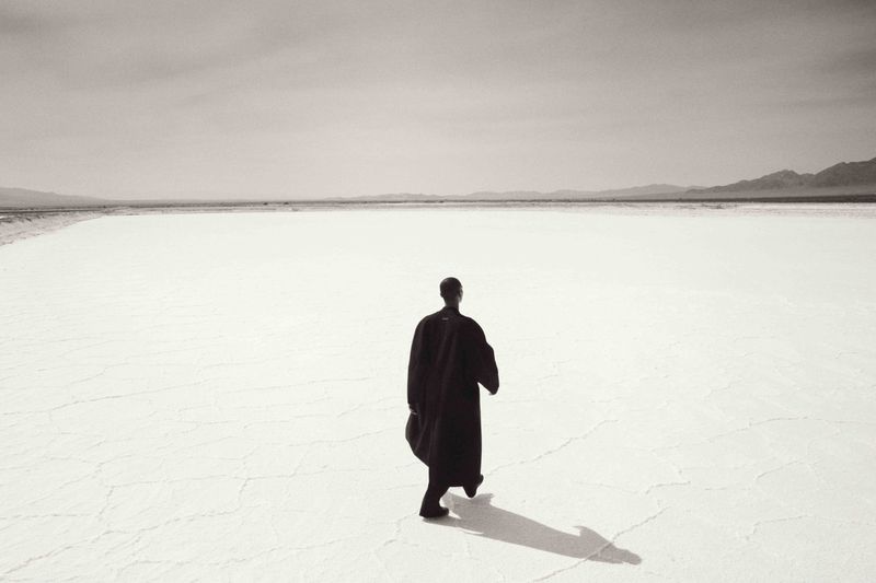Back view of a person walking in a dark robe across a bright white desert expanse toward distant hills, with a strong cast shadow.