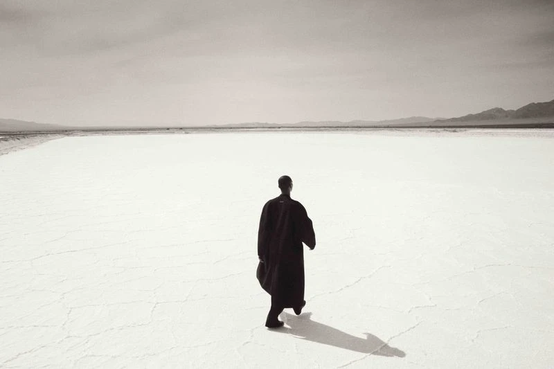 Back view of a person walking in a dark robe across a bright white desert expanse toward distant hills, with a strong cast shadow.