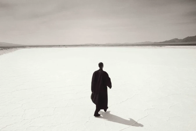 Back view of a person walking in a dark robe across a bright white desert expanse toward distant hills, with a strong cast shadow.
