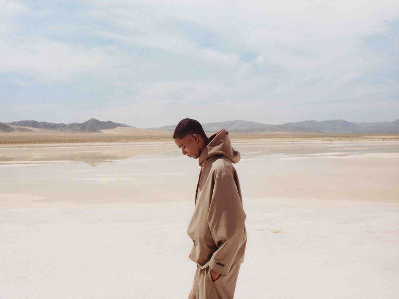 A person stands on a beige, reflective lakebed with mountains in the background under a light blue sky with wispy clouds. 
