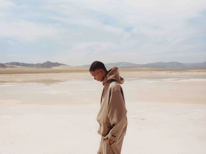 A person stands on a beige, reflective lakebed with mountains in the background under a light blue sky with wispy clouds. 