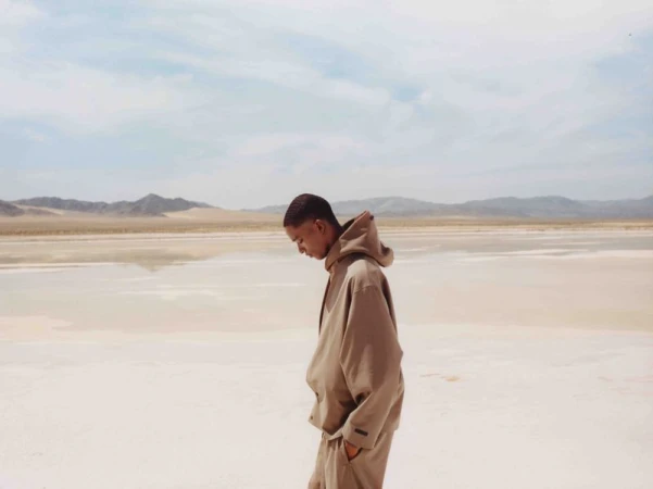 A person stands on a beige, reflective lakebed with mountains in the background under a light blue sky with wispy clouds. 