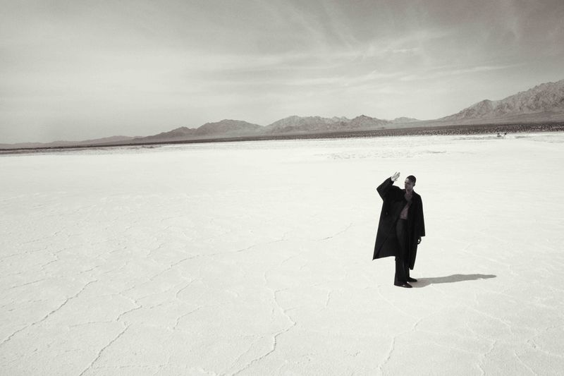 Monochrome portrait of an adult male in an overcoat standing on a cracked plain with distant mountains under a cloudy sky.