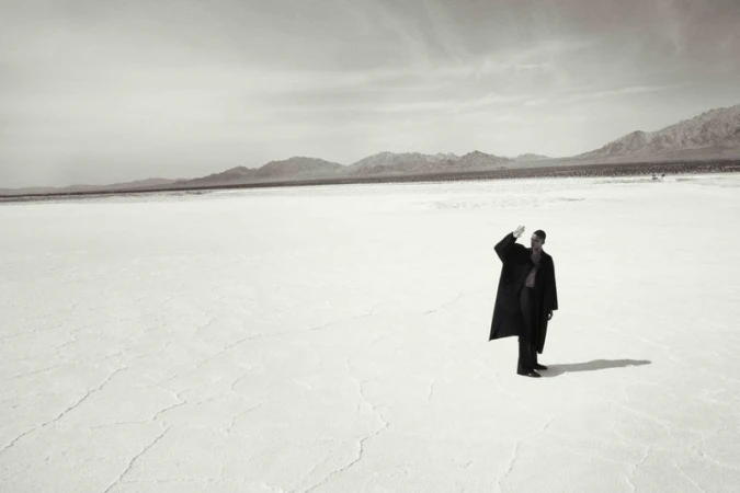 Monochrome portrait of an adult male in an overcoat standing on a cracked plain with distant mountains under a cloudy sky.