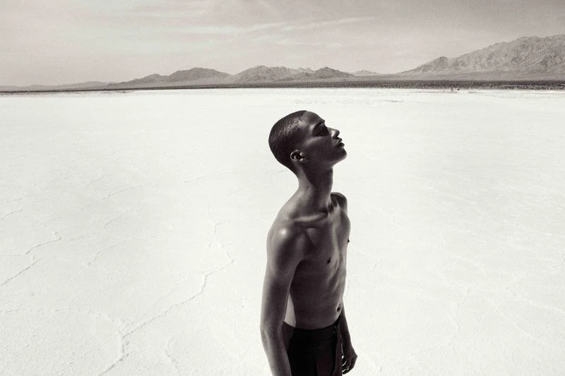 Monochrome portrait of a Black man looking up in an expansive white landscape with mountains on the horizon, set under a bright sky.