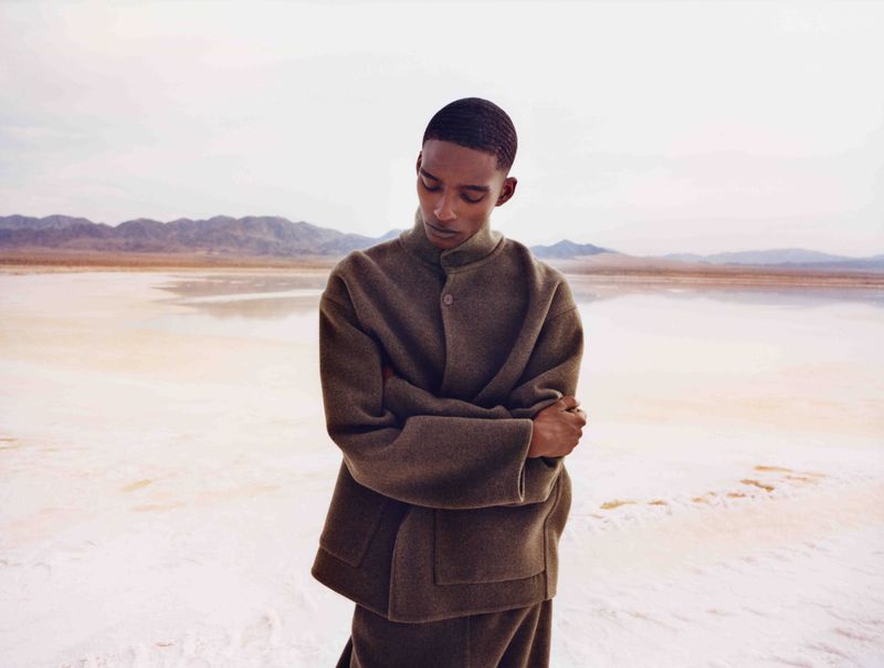 An adult male poses in a relaxed position in a desert landscape with salt flats. He is wearing an olive-green, wool-blend jacket.
