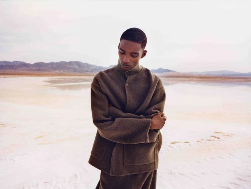 An adult male poses in a relaxed position in a desert landscape with salt flats. He is wearing an olive-green, wool-blend jacket.