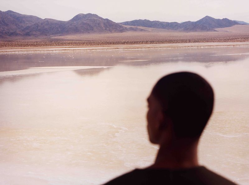 A person stands before a vast, reflective expanse resembling a salt flat, with distant mountains under an expansive sky.
