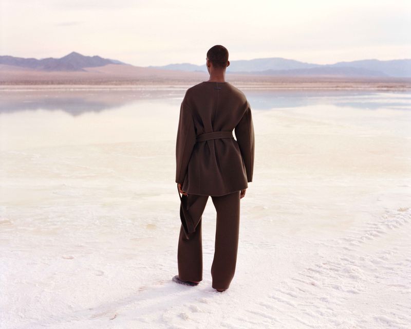 Person in a brown outfit standing on a white surface with mountains in the background, facing away from the camera.