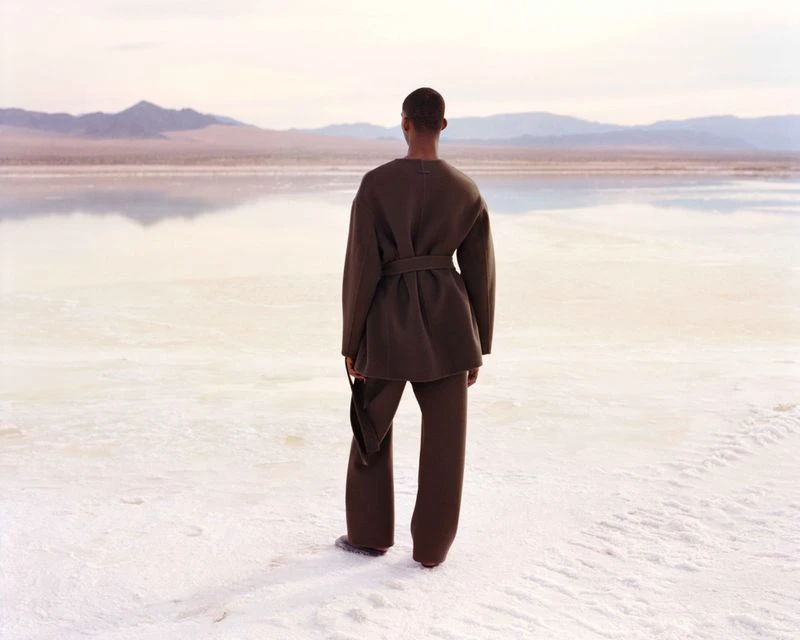 Person in a brown outfit standing on a white surface with mountains in the background, facing away from the camera.