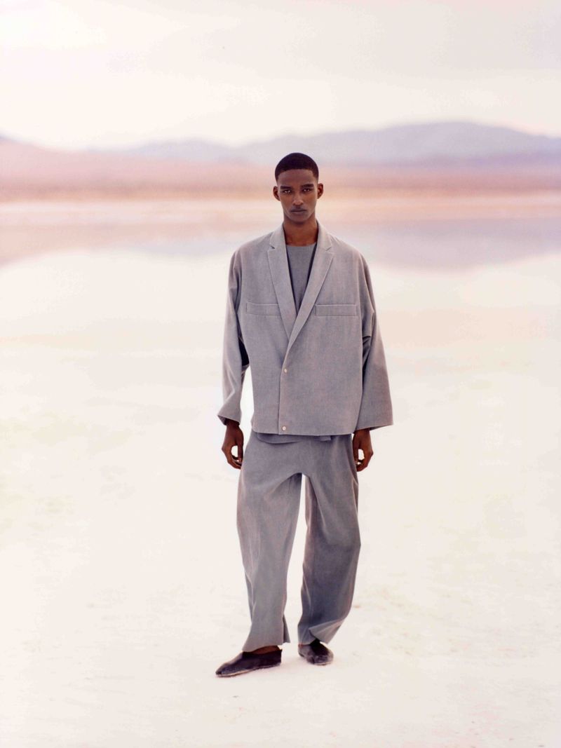 A lone adult male is standing in a spacious, brightly lit outdoor area, clad in grey blazer and matching trousers, with a distant backdrop.