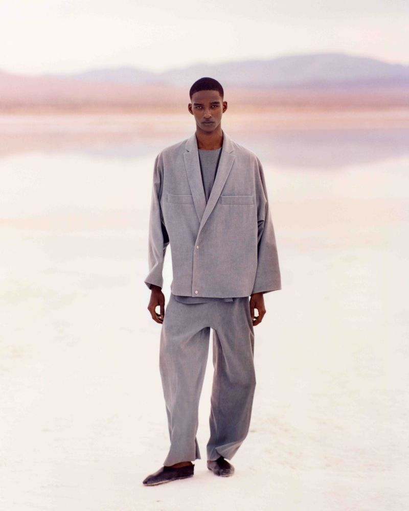 A full shot of a Black male model in a grey, oversized suit stands in a pale landscape with distant, blurry mountain shapes under muted light.