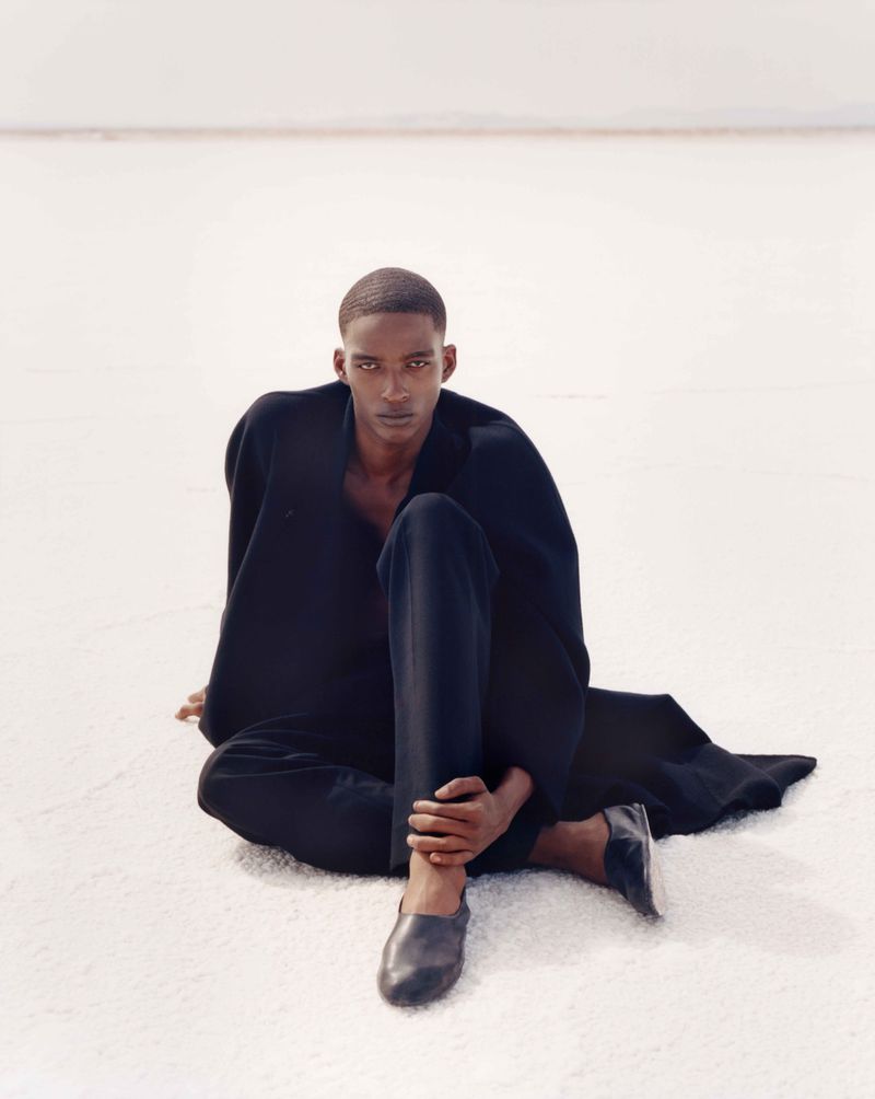 A Black male model in a dark outfit sits on a light-colored surface in a minimalist, high-key fashion shot with shallow depth of field.