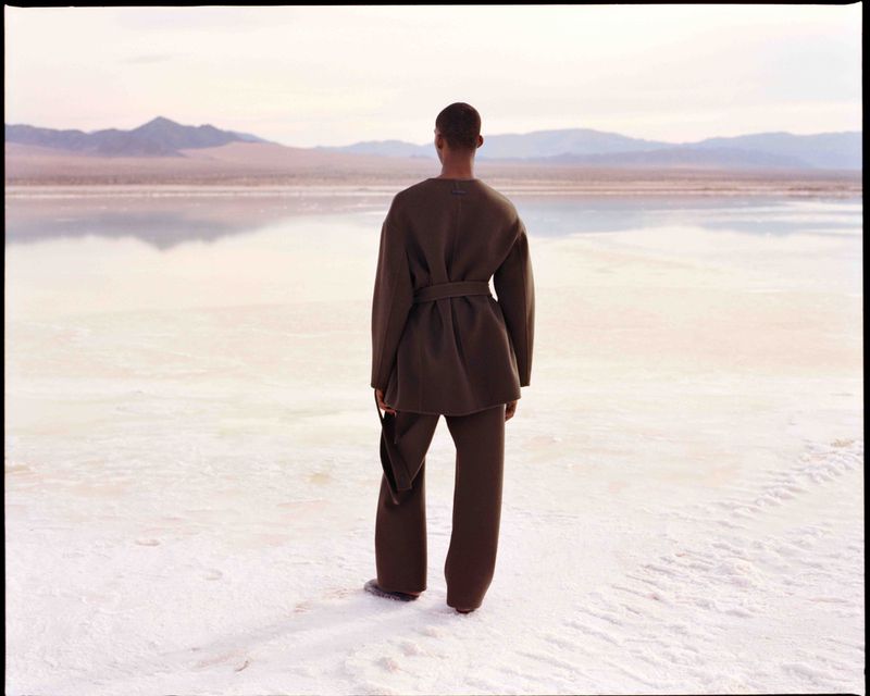 A person stands in a brown outfit on a white expanse overlooking a lake and mountains in the distance, creating a tranquil scene.