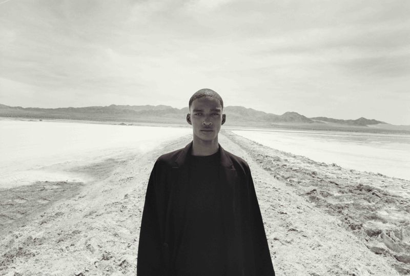 Monochrome portrait of a young adult stands on a textured dirt road, a vast salt flat and distant mountains under a cloudy sky stretch behind.
