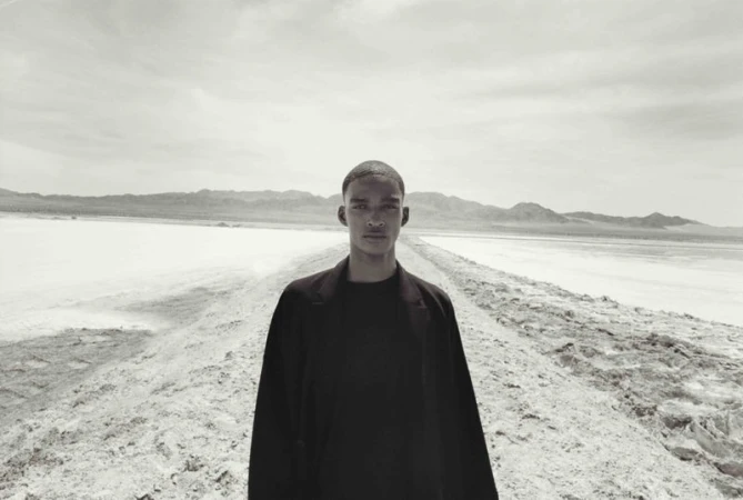 Monochrome portrait of a young adult stands on a textured dirt road, a vast salt flat and distant mountains under a cloudy sky stretch behind.