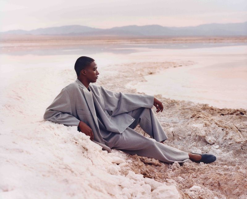 A dark-skinned male model wearing a grey suit reclines on a textured white surface near a pink lake with mountains in the background.