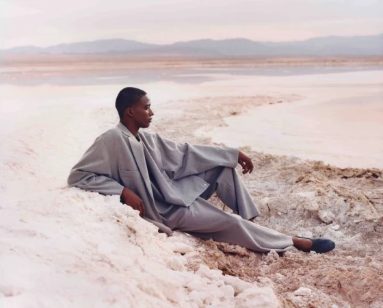 A dark-skinned male model wearing a grey suit reclines on a textured white surface near a pink lake with mountains in the background.