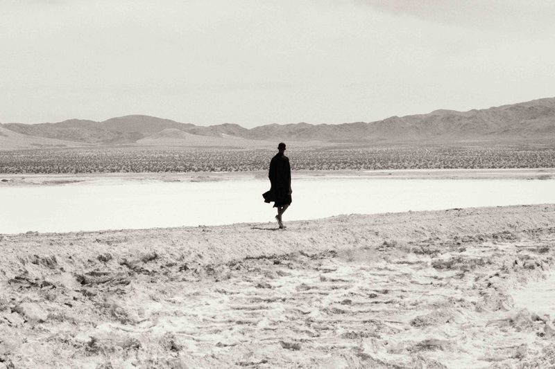 Monochrome photograph of an adult walking barefoot on a sandbar in a desert landscape, with mountains in the background on a sunny day.
