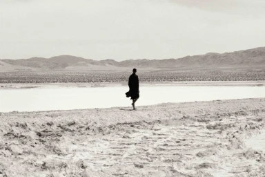 Model walking across a pale desert landscape wearing a long dark coat with distant mountains.