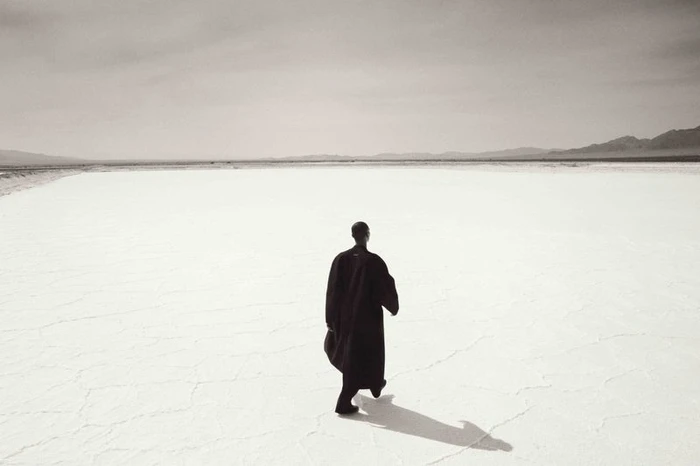 Model in a long black coat walking away across an expansive white salt flat under a pale sky.