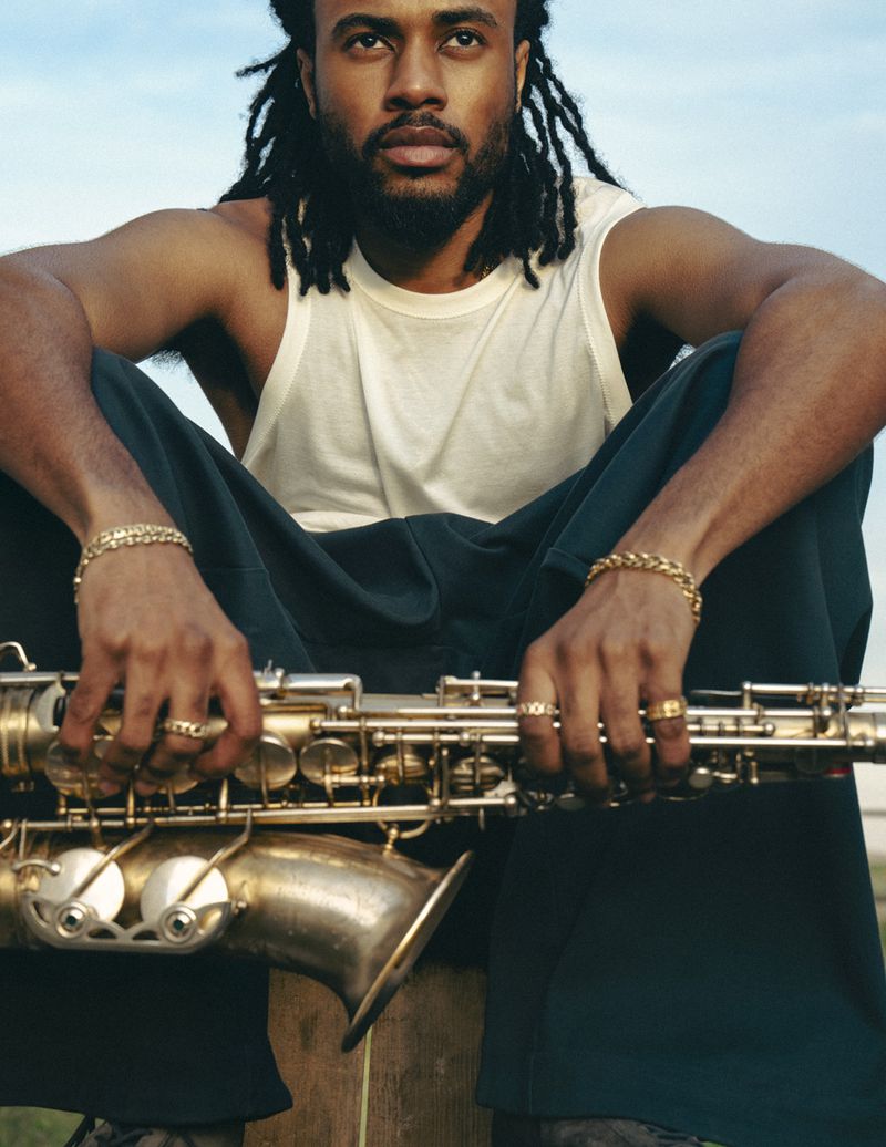 Portrait of Black man with dreadlocks sitting outdoors holding a saxophone. He wears a white tank top and dark pants.