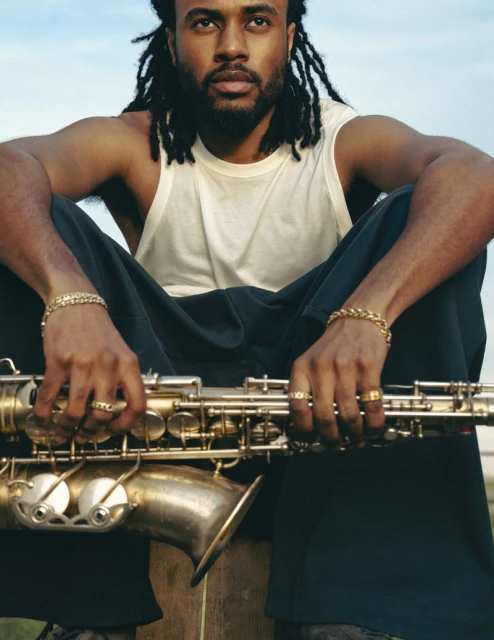 Portrait of Black man with dreadlocks sitting outdoors holding a saxophone. He wears a white tank top and dark pants.