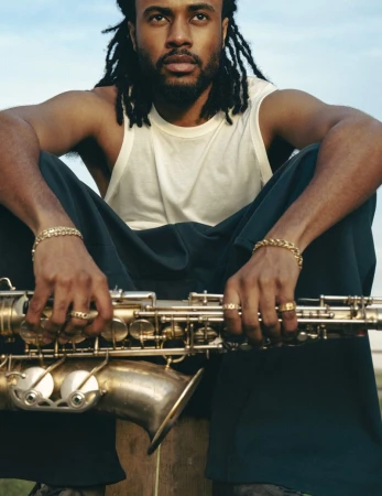 Portrait of Black man with dreadlocks sitting outdoors holding a saxophone. He wears a white tank top and dark pants.